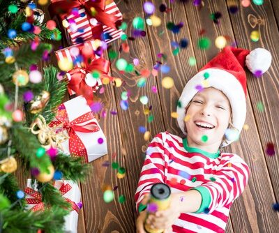 a young boy in red and white striped pajamas is laying on his back under a colorfully decorate Christmas tree, he is wearing a red Santa hat and is holding a party popper that is releasing confetti all around him