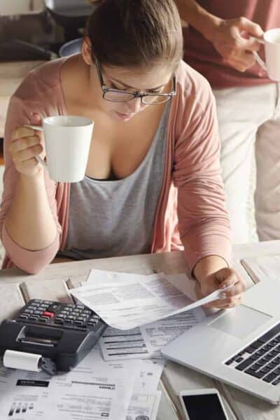 Top view of serious young woman wearing glasses managing family budget, calculating domestic expenses, paying utility bills online, using laptop pc and calculator while drinking coffee in kitchen