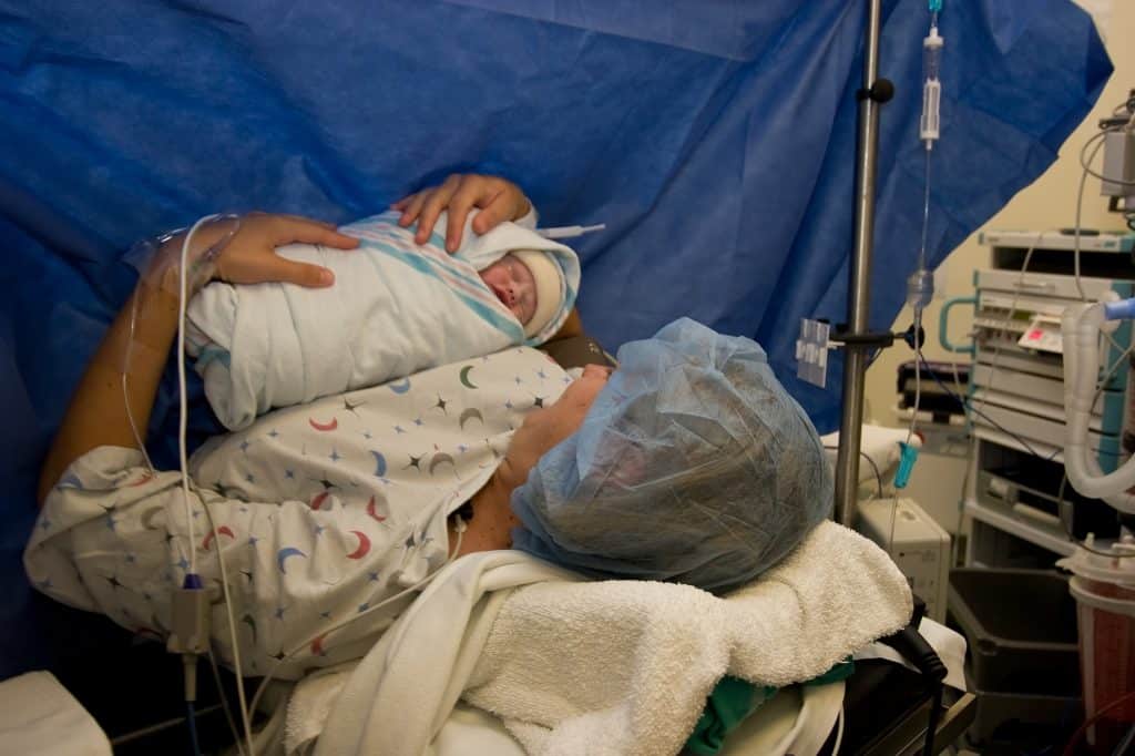 a new mom is holding her newborn baby; mom is in a surgical cap and surgical gown, she is still on the operating table after her c section